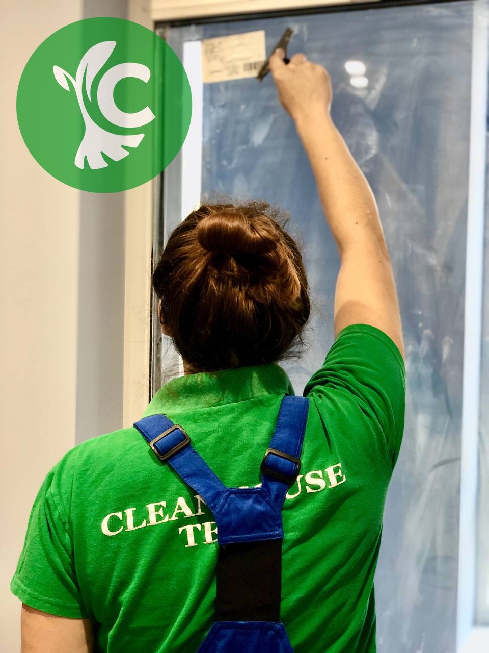 Pracownica firmy sprzątającej w zielonej koszulce z logo, czyści okno za pomocą skrobaczki, widok z tyłu.