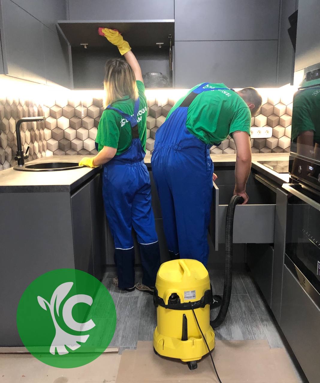 Dwoje pracowników w niebieskich kombinezonach i zielonych koszulkach, sprzątają szarą kuchnię z heksagonalnymi płytkami. Jeden czyści szafkę, drugi odkurza otwartą szufladę, obok stoi żółty odkurzacz.
