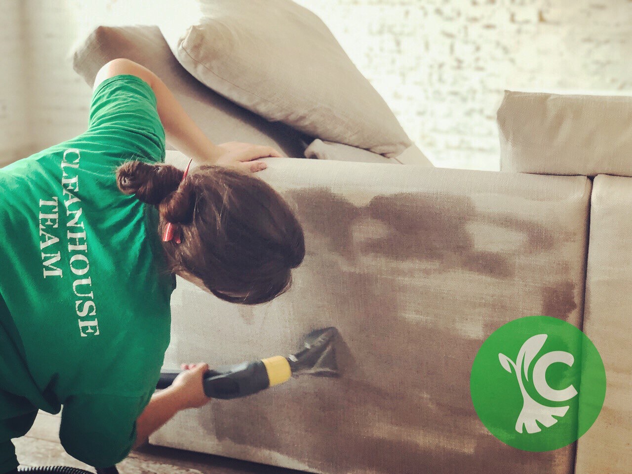 Kobieta w zielonej koszulce z napisem 'CLEANHOUSE TEAM' czyści jasną tapicerkę sofy za pomocą odkurzacza piorącego; widoczne mokre plamy na materiale i logo firmy w prawym dolnym rogu.