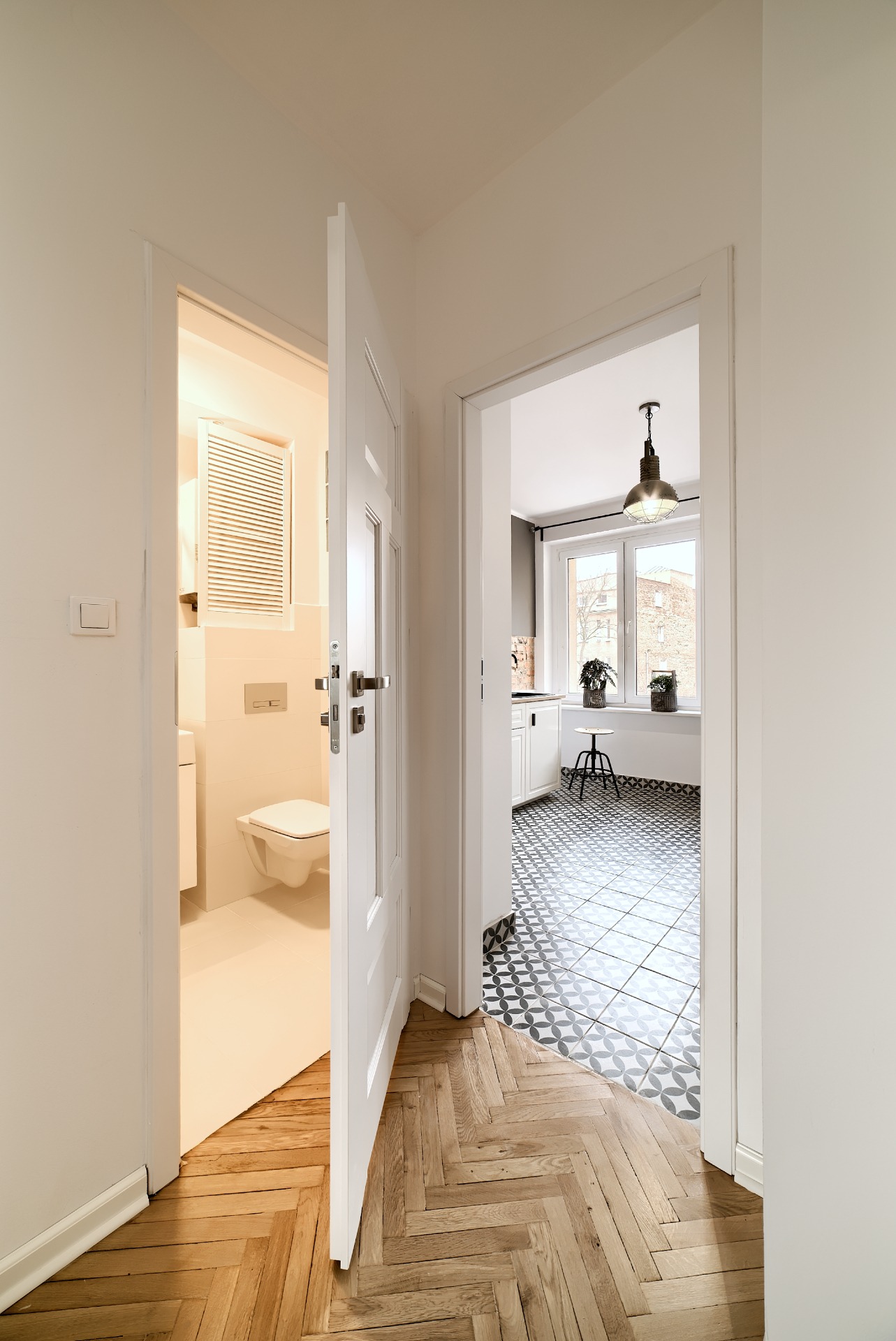 Widok na wyremontowaną łazienkę i kuchnię z korytarza z parkietem w jodełkę, białe drzwi otwarte, wzorzyste płytki podłogowe w kuchni, minimalistyczny wystrój.