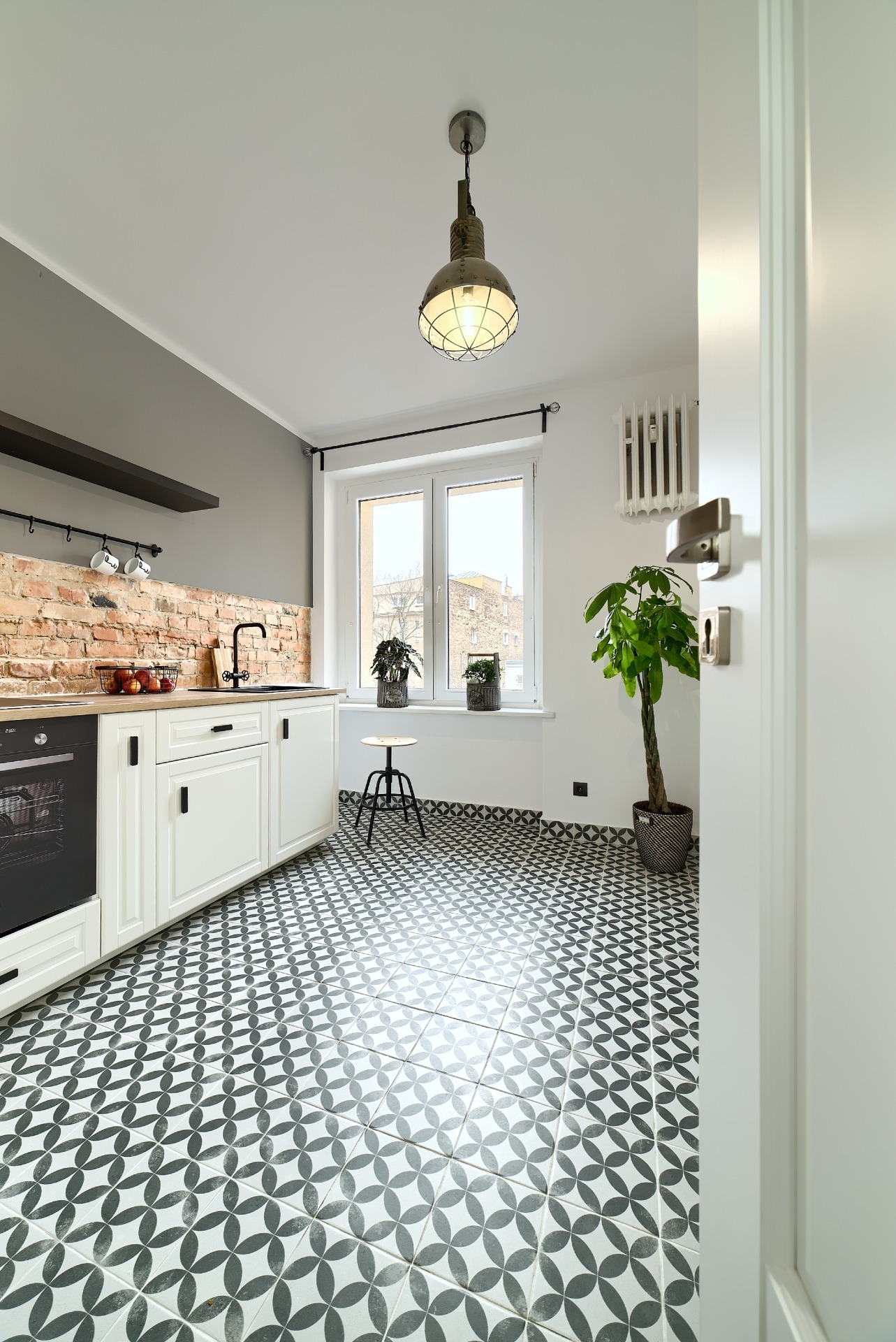 Jasna kuchnia z białymi szafkami, ceglaną ścianą nad blatem, czarną armaturą i wzorzystą podłogą w stylu retro, doświetlona lampą w industrialnym stylu.