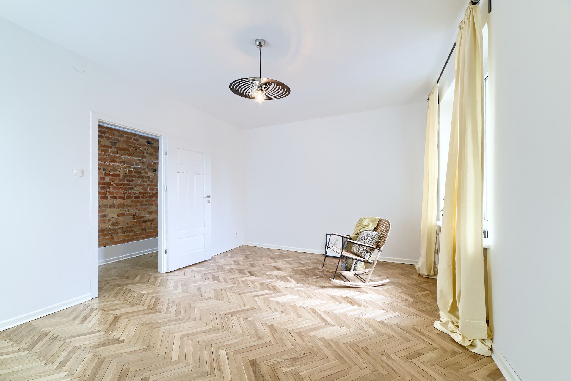 Jasne, puste pomieszczenie z parkietem w jodełkę, otwartymi białymi drzwiami ukazującymi ceglaną ścianę, designerską lampą sufitową i wiklinowym fotelem bujanym z poduszką i pledem przy oknie...