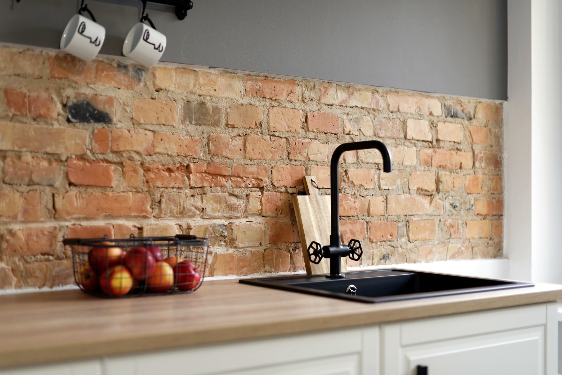 Kuchnia w stylu loft: czarny zlew z baterią retro na tle ceglanej ściany, koszyk z jabłkami na blacie, dwie filiżanki wiszące nad ścianą.