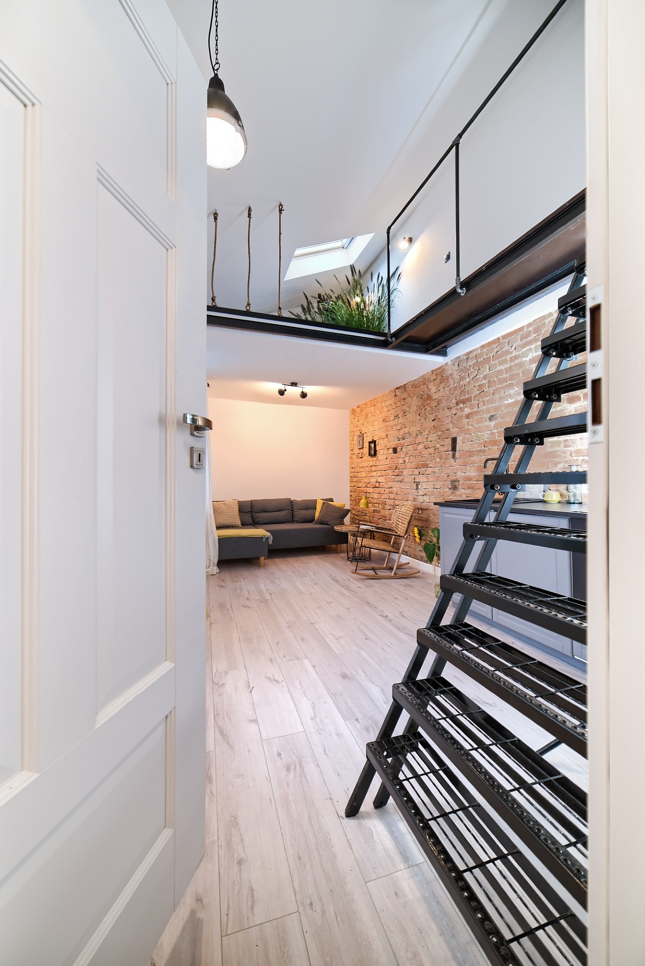 Otwarty widok na salon z antresolą, ukazujący metalowe schody drabinkowe, fragment ceglanej ściany i roślinność na górze, drewniana podłoga i szara sofa w tle, widziane przez otwarte białe drzwi.