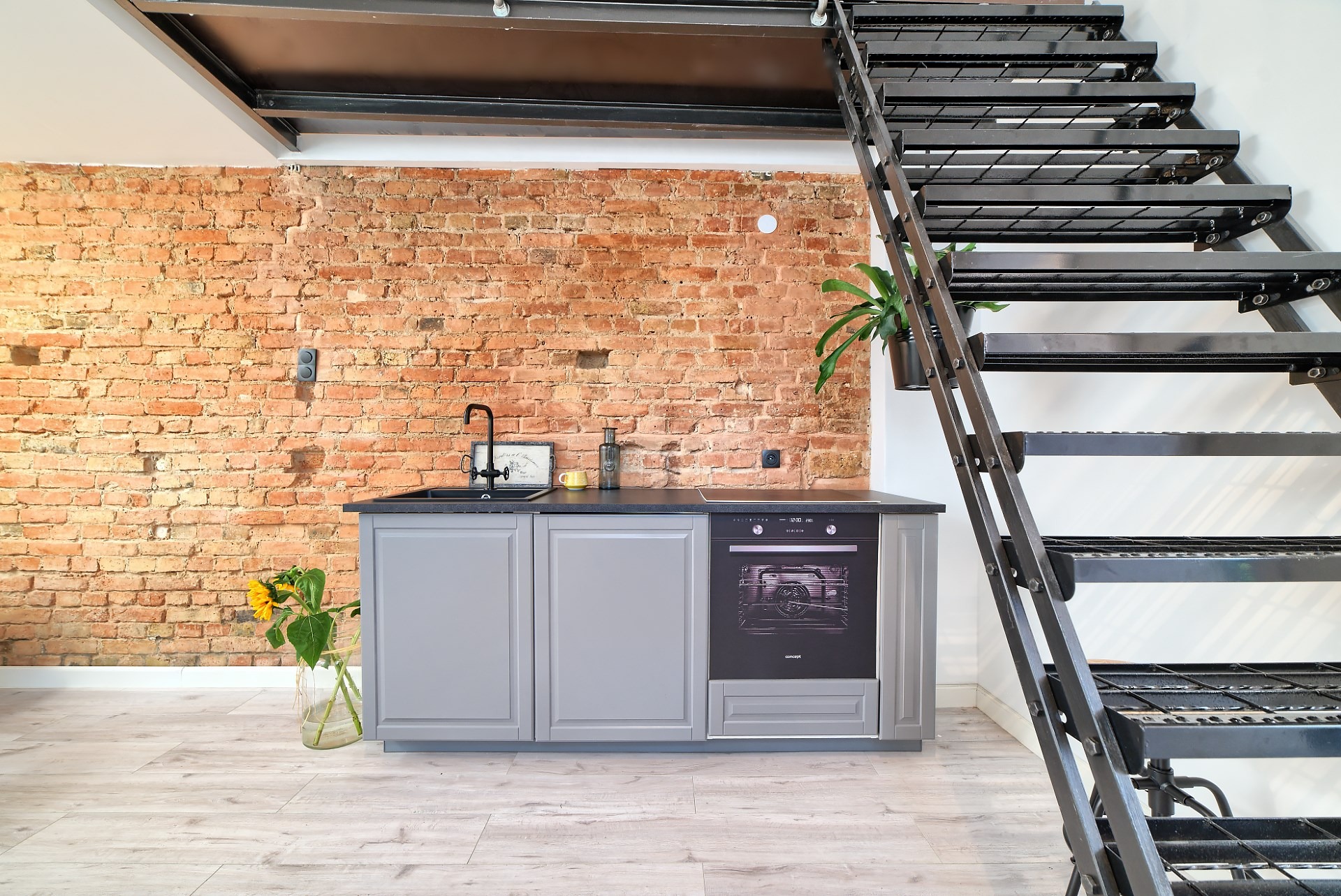 Kompaktowa kuchnia z szarymi szafkami i czarnym blatem na tle ceglanej ściany, z metalowymi schodami prowadzącymi na antresolę, oświetlona naturalnym światłem.