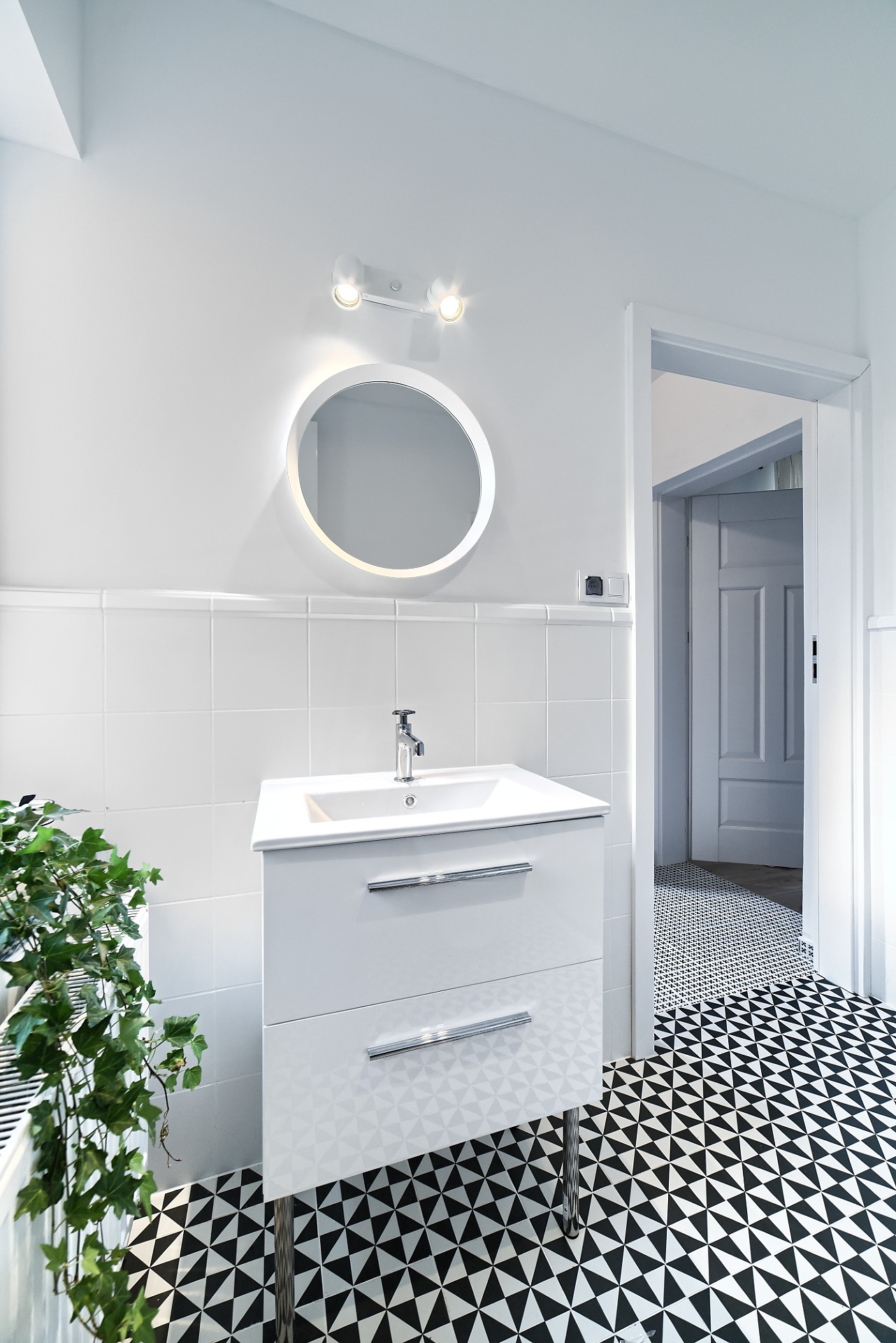 Nowoczesna biała łazienka z geometryczną czarno-białą podłogą, okrągłym podświetlanym lustrem i rośliną doniczkową obok umywalki z szafką.