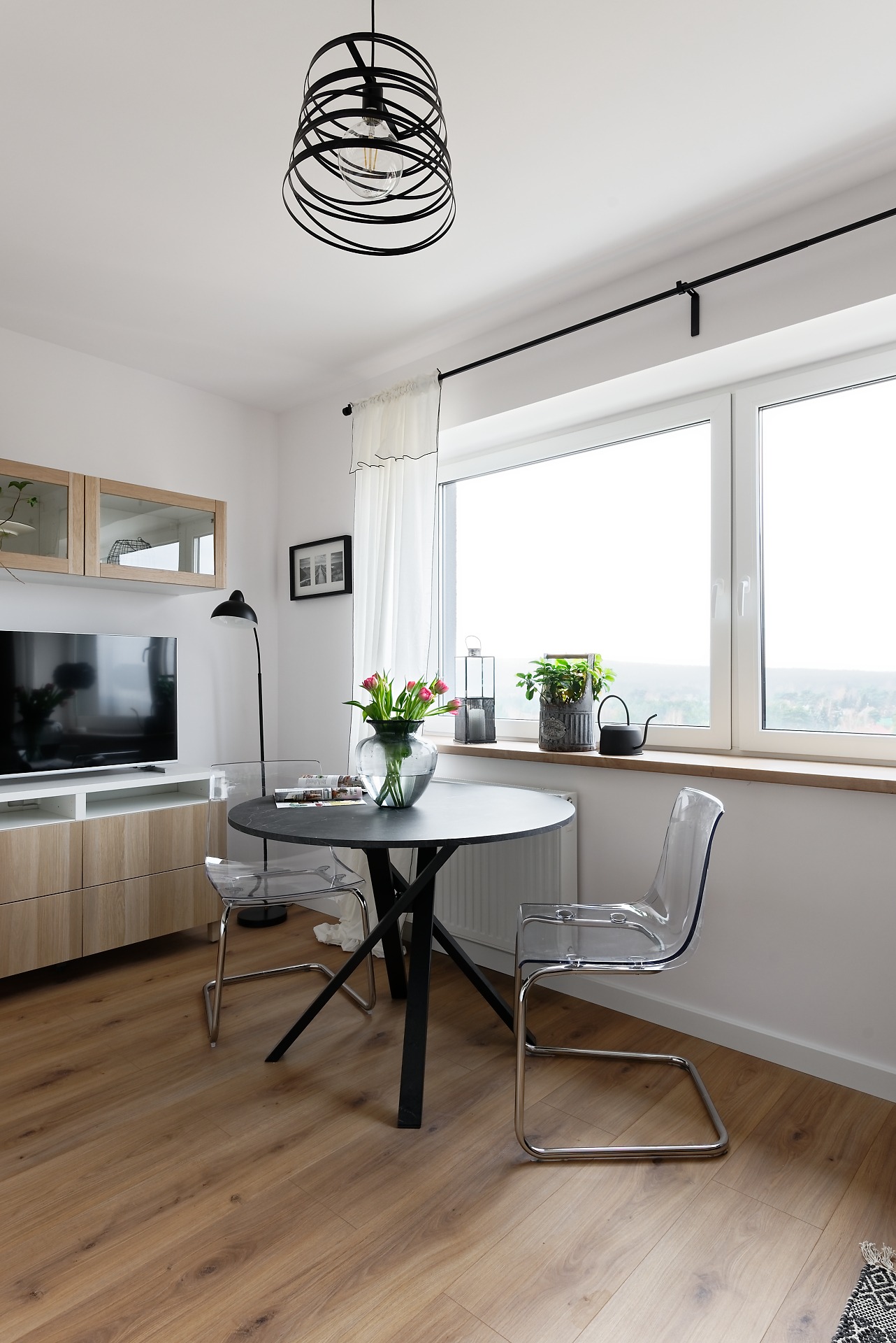 Jasne wnętrze z okrągłym czarnym stołem, przezroczystymi krzesłami, drewnianą podłogą i geometryczną lampą wiszącą.