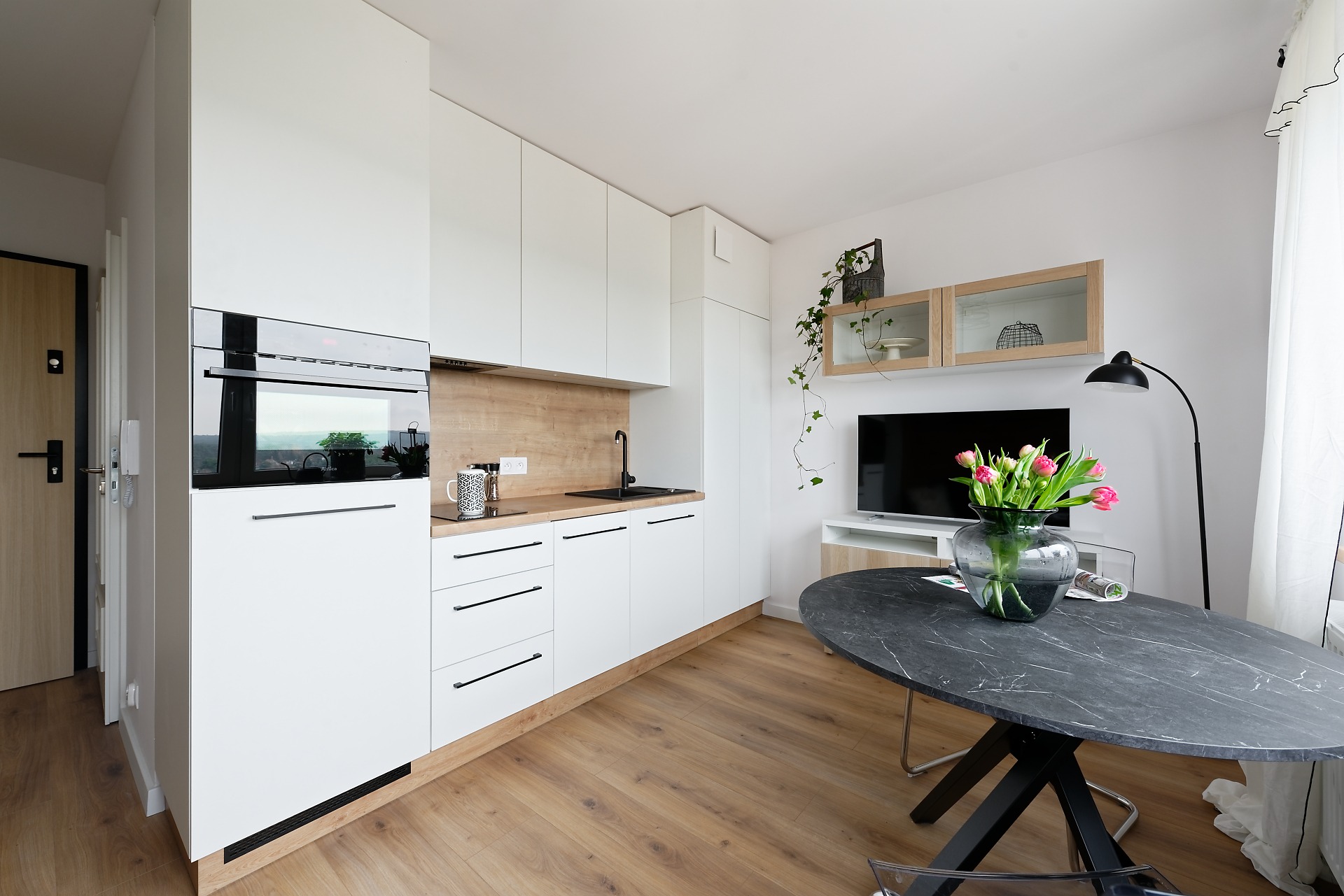 Jasne wnętrze mieszkania z białą zabudową kuchenną, drewnianym blatem i podłogą, czarnym zlewem i marmurowym stołem z tulipanami w wazonie.