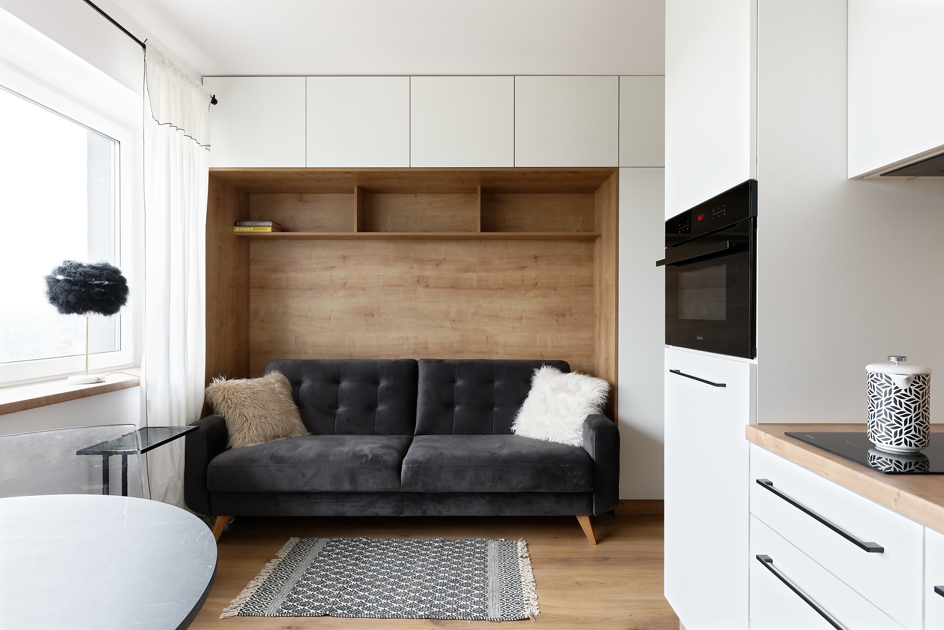 Nowoczesne wnętrze: szara sofa z puszystymi poduszkami w drewnianej wnęce obok białej zabudowy kuchennej z czarnym piekarnikiem i płytą indukcyjną.