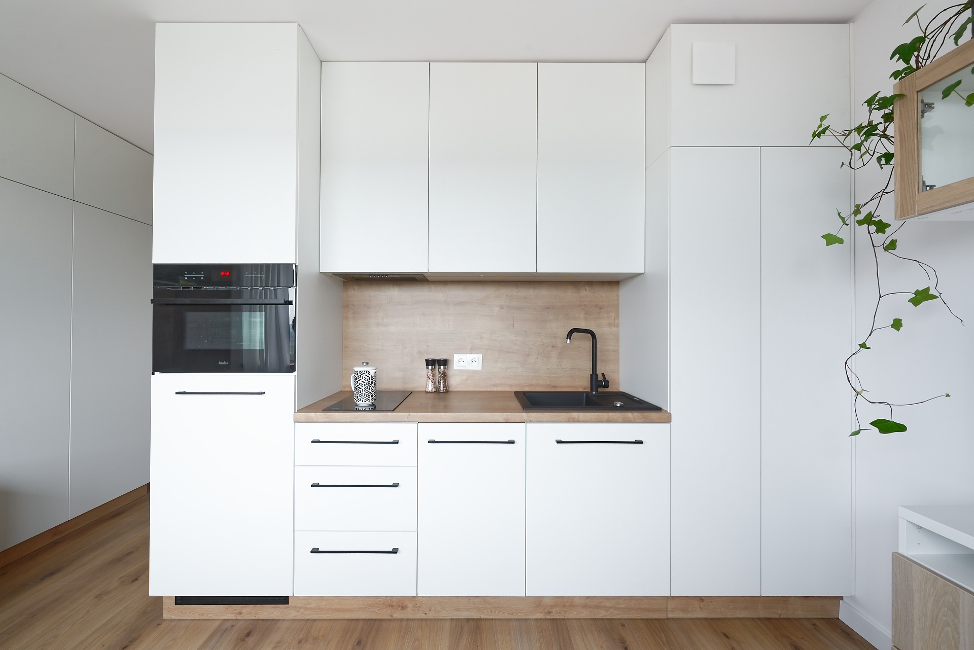 Nowoczesna, minimalistyczna kuchnia z białymi frontami szafek i drewnianym blatem, czarny zlew i bateria, sprzęt AGD w zabudowie, akcent roślinny w postaci pnącza na ścianie.