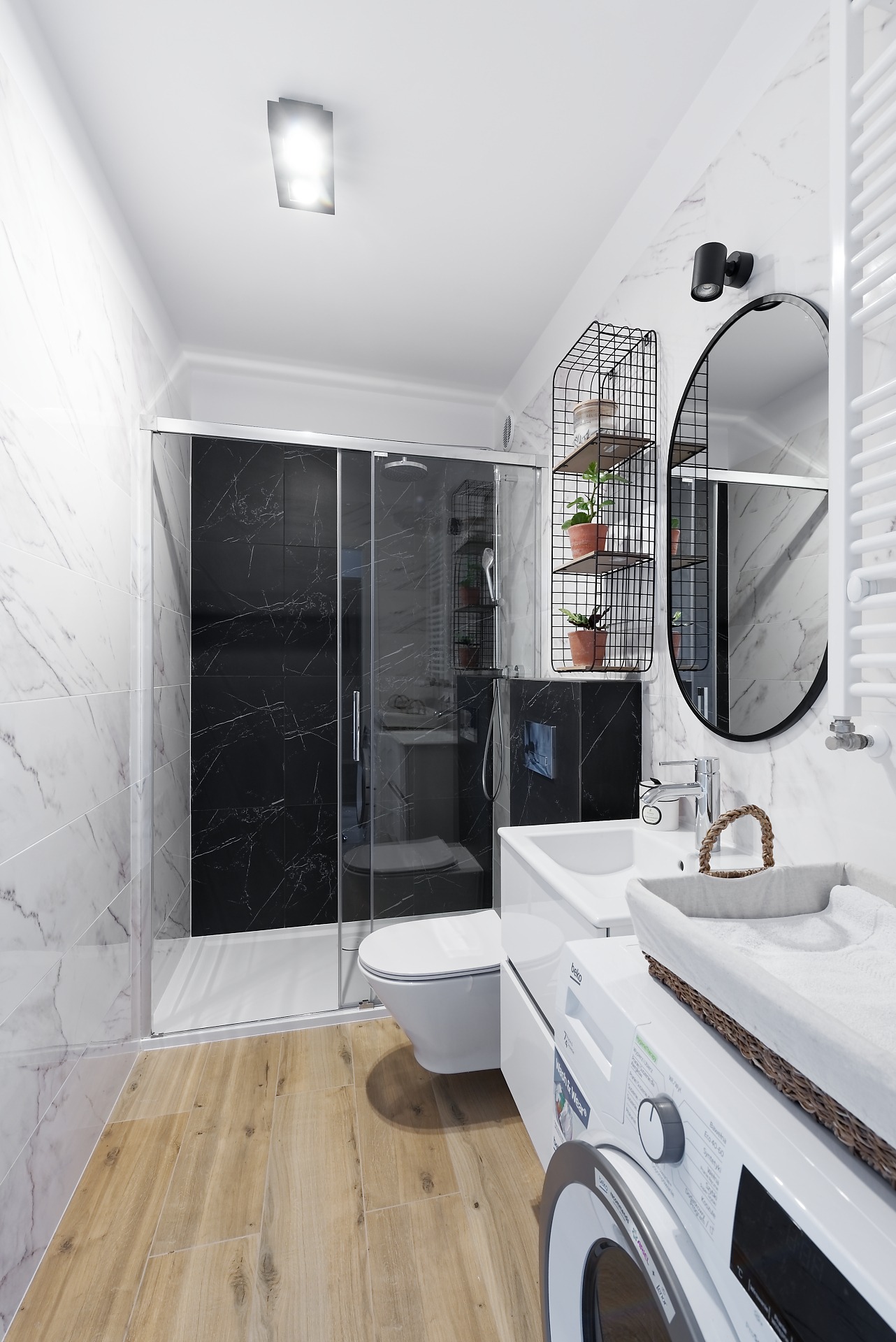 Nowoczesna łazienka z kabiną prysznicową z czarnymi płytkami imitującymi marmur, białymi płytkami z marmurowym wzorem na ścianach, drewnopodobną podłogą, umywalką z szafką, okrągłym lustrem...