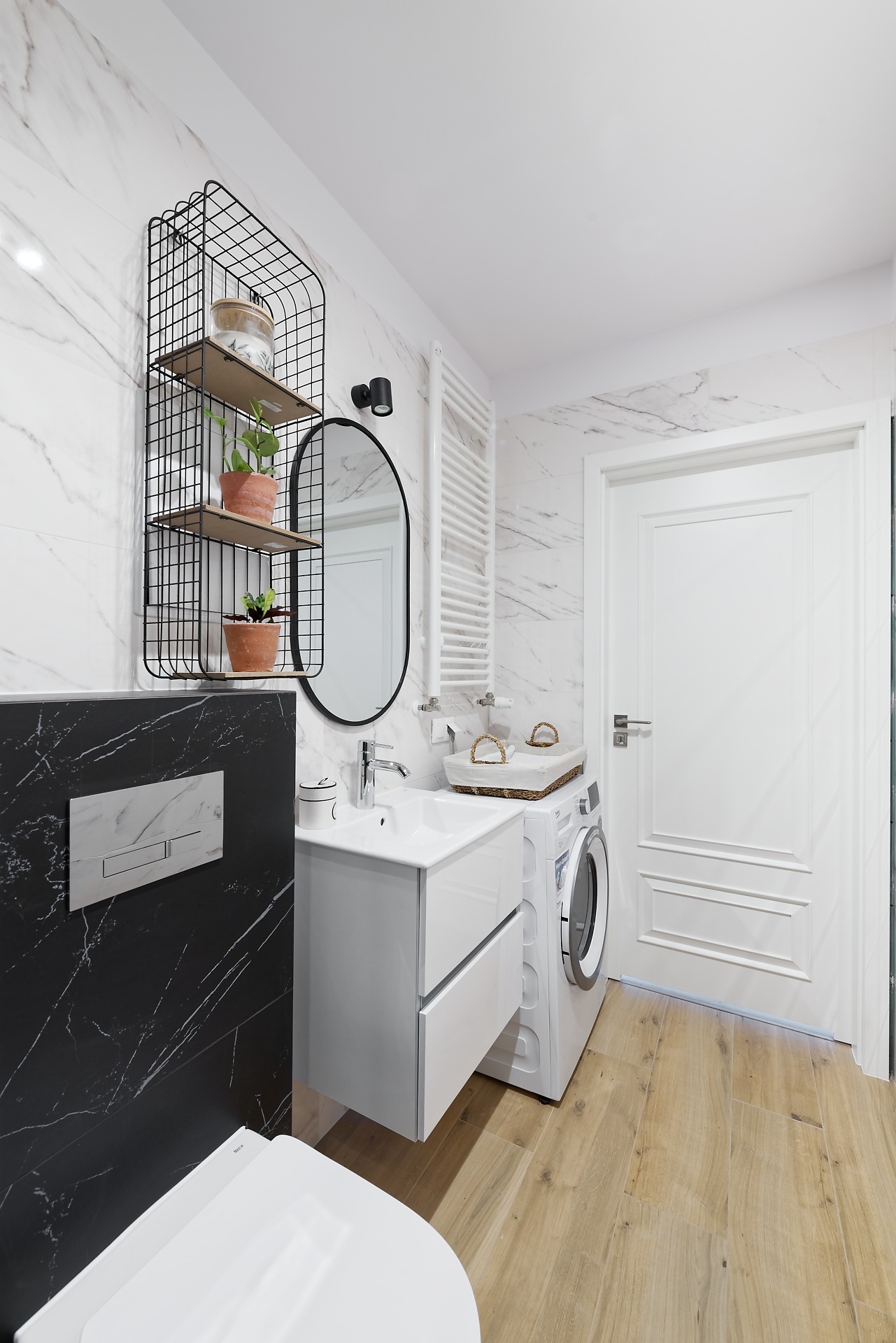 Nowoczesna łazienka z marmurowymi płytkami, białymi meblami, wiszącą półką z roślinami i pralką.