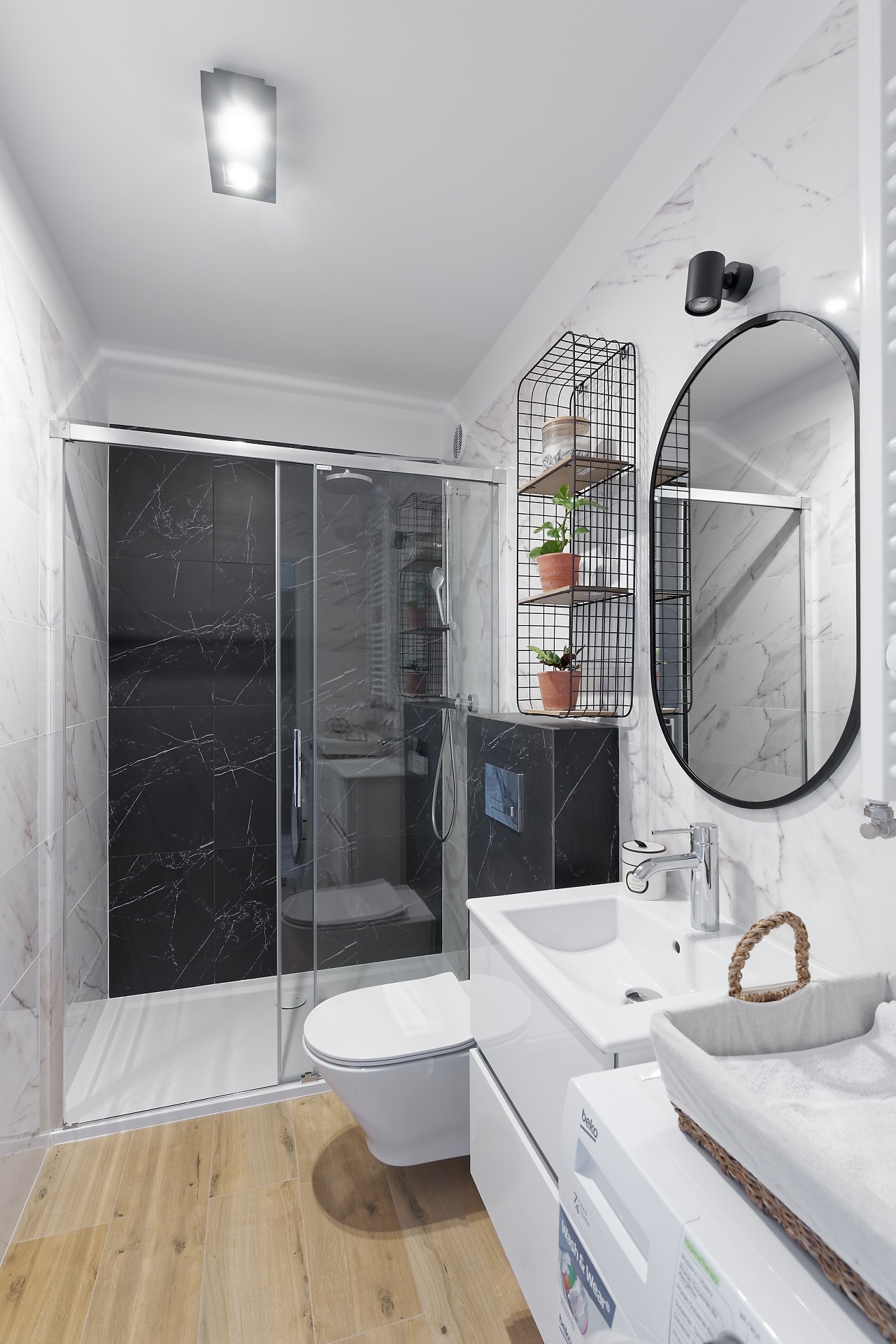 Nowoczesna łazienka z kabiną prysznicową typu walk-in z czarnymi płytkami imitującymi marmur, wiszącą toaletą, umywalką z szafką, owalnym lustrem w czarnej ramie i drewnopodobną podłogą, oświetlona...