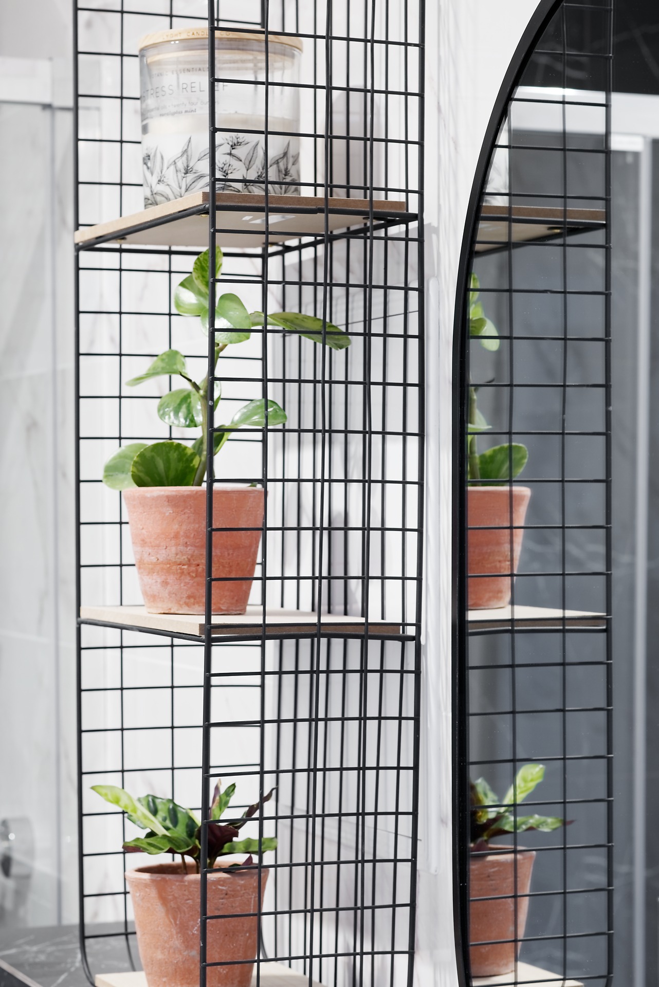 Metalowy regał z drewnianymi półkami w łazience, na którym stoją dwie doniczki z roślinami i świeca, odbicie rośliny w lustrze.