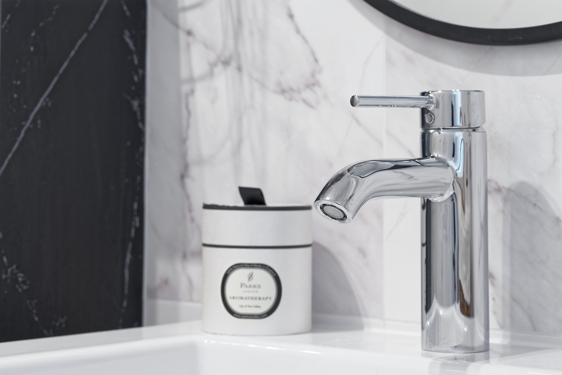 Błyszcząca chromowana bateria umywalkowa z cylindrycznym dyfuzorem, obok aromatyczna świeca w białym pojemniku, na tle marmurowej ściany w odcieniach bieli i szarości.