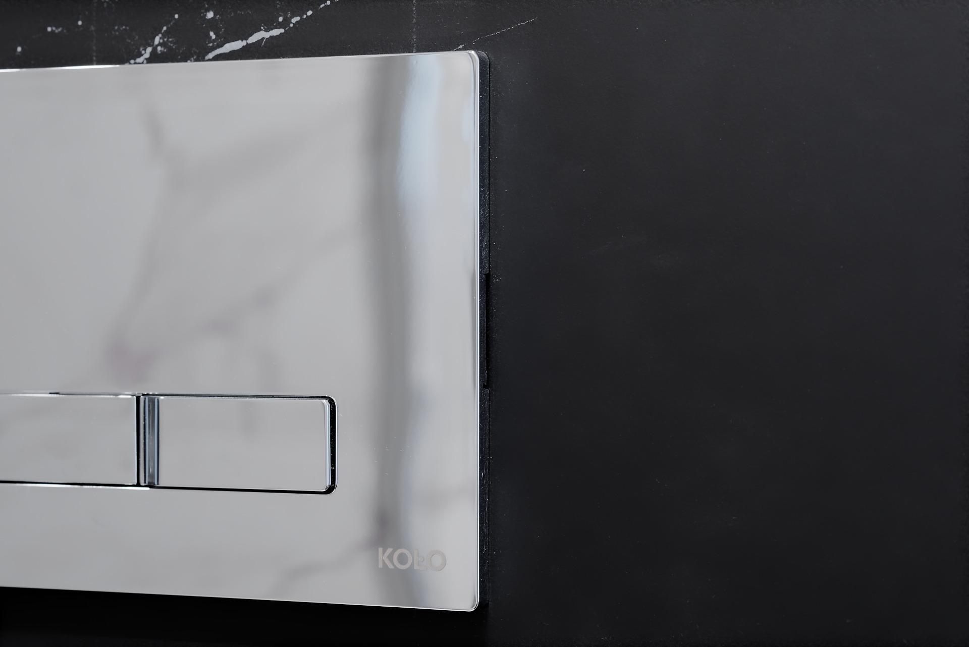 Błyszczący, chromowany przycisk spłuczki KOLO na tle czarnej, marmurowej ściany w łazience.