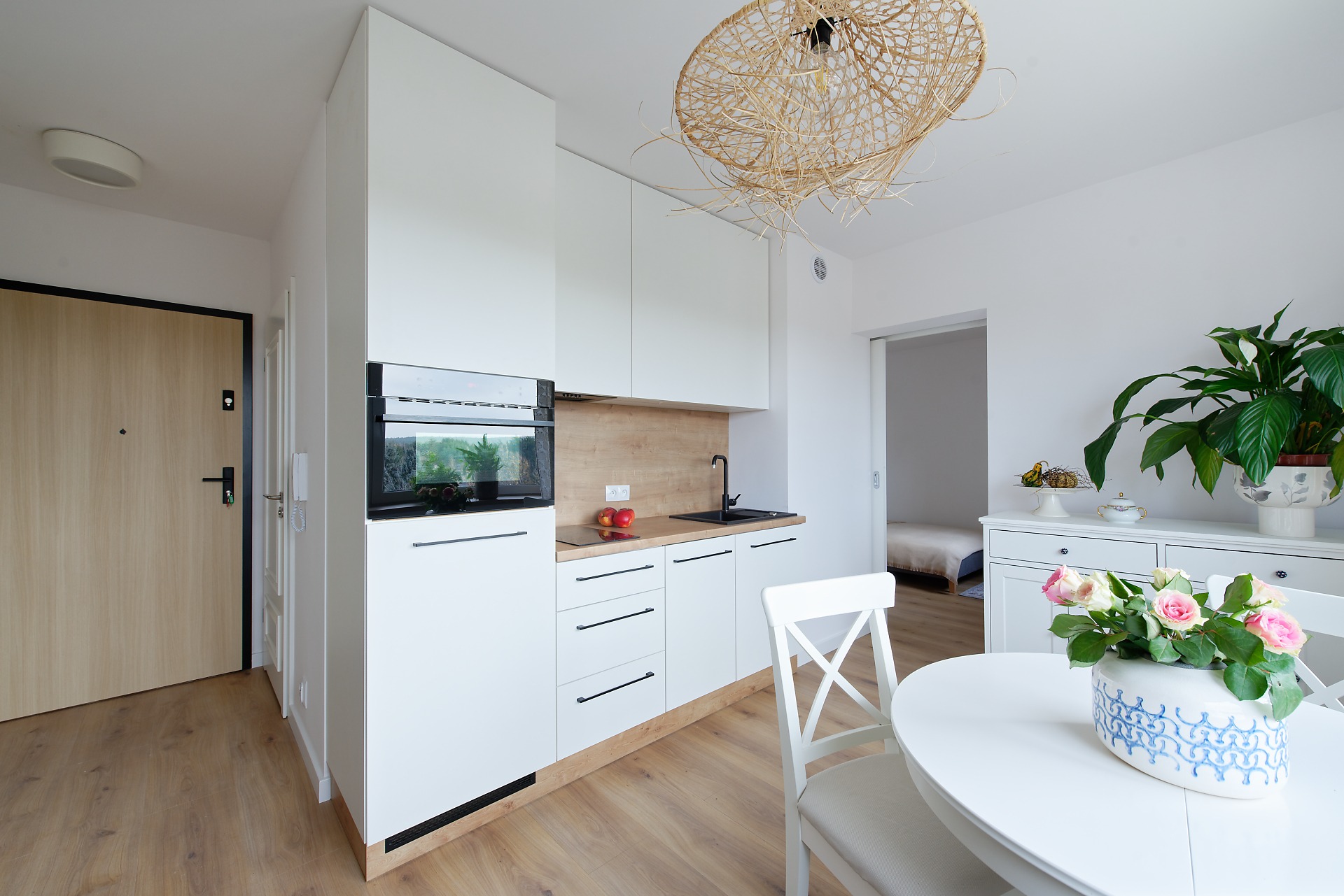 Jasna kuchnia z białymi meblami, drewnianym blatem i plecionym żyrandolem, widok na stół z kwiatami i drzwi wejściowe