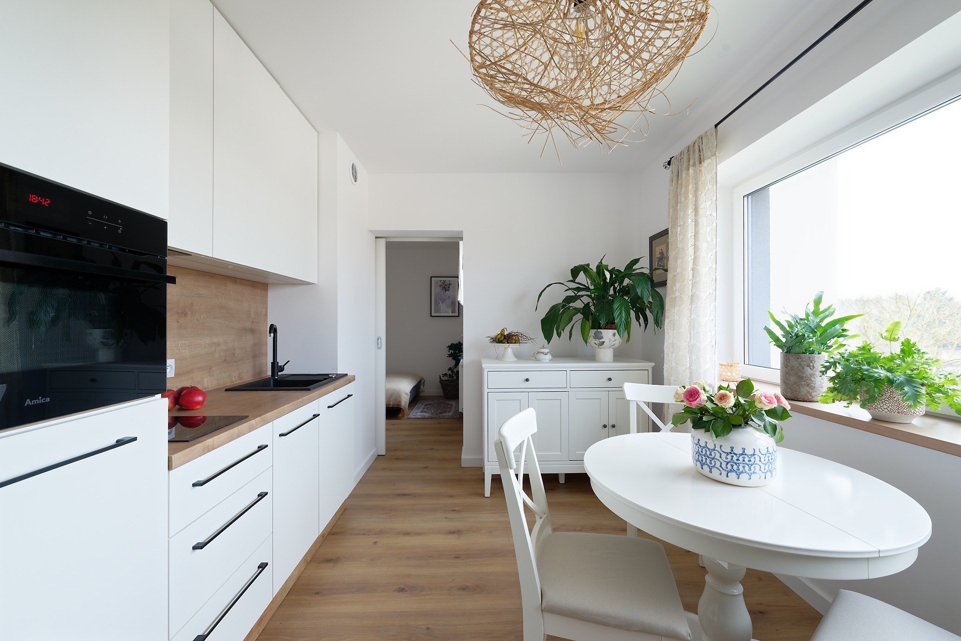 Jasna kuchnia w stylu skandynawskim z drewnianymi akcentami, okrągłym stołem i roślinami w doniczkach przy oknie.