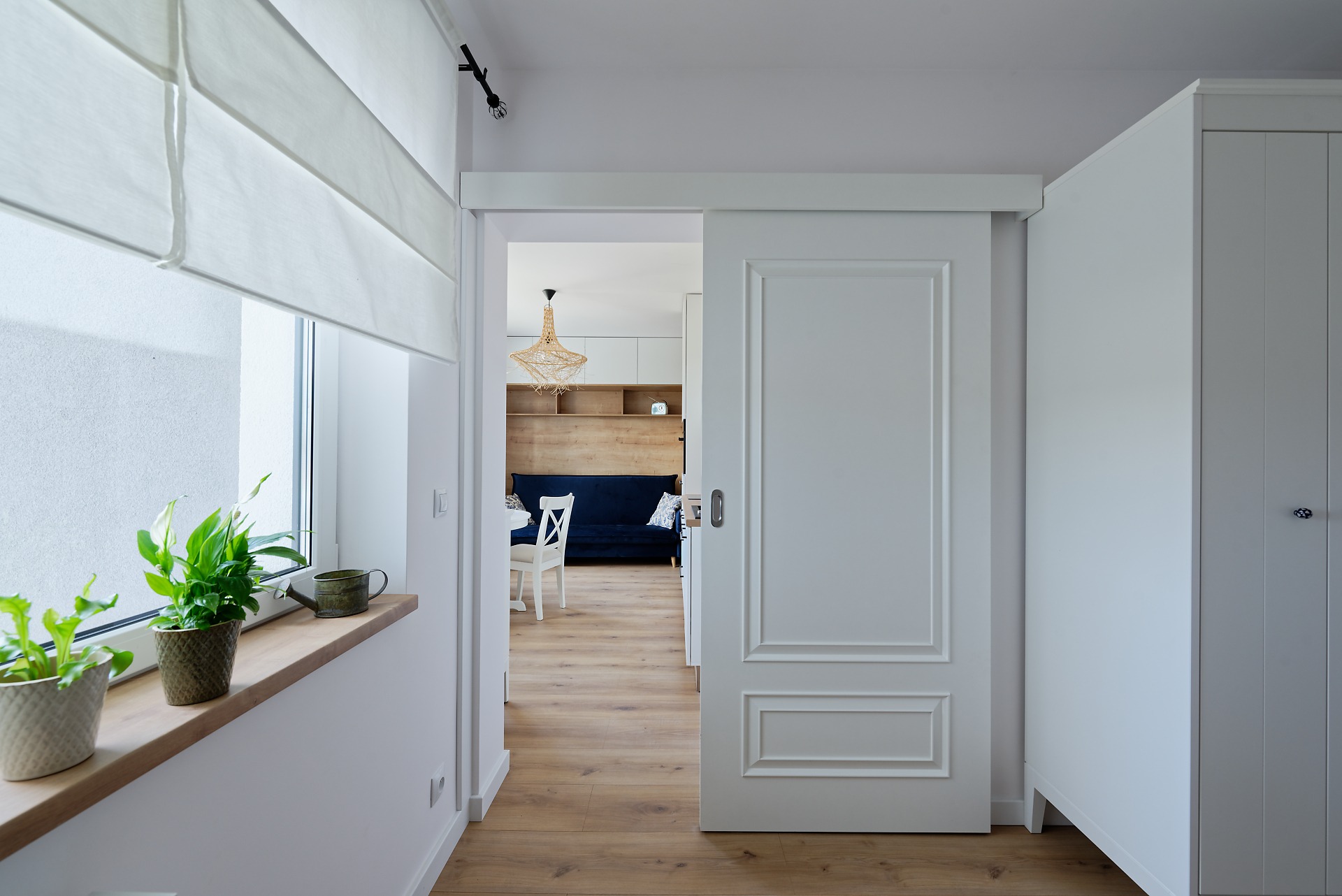 Nowoczesne wnętrze mieszkania z przesuwnymi białymi drzwiami, drewnianą podłogą i roślinami doniczkowymi na parapecie okiennym.