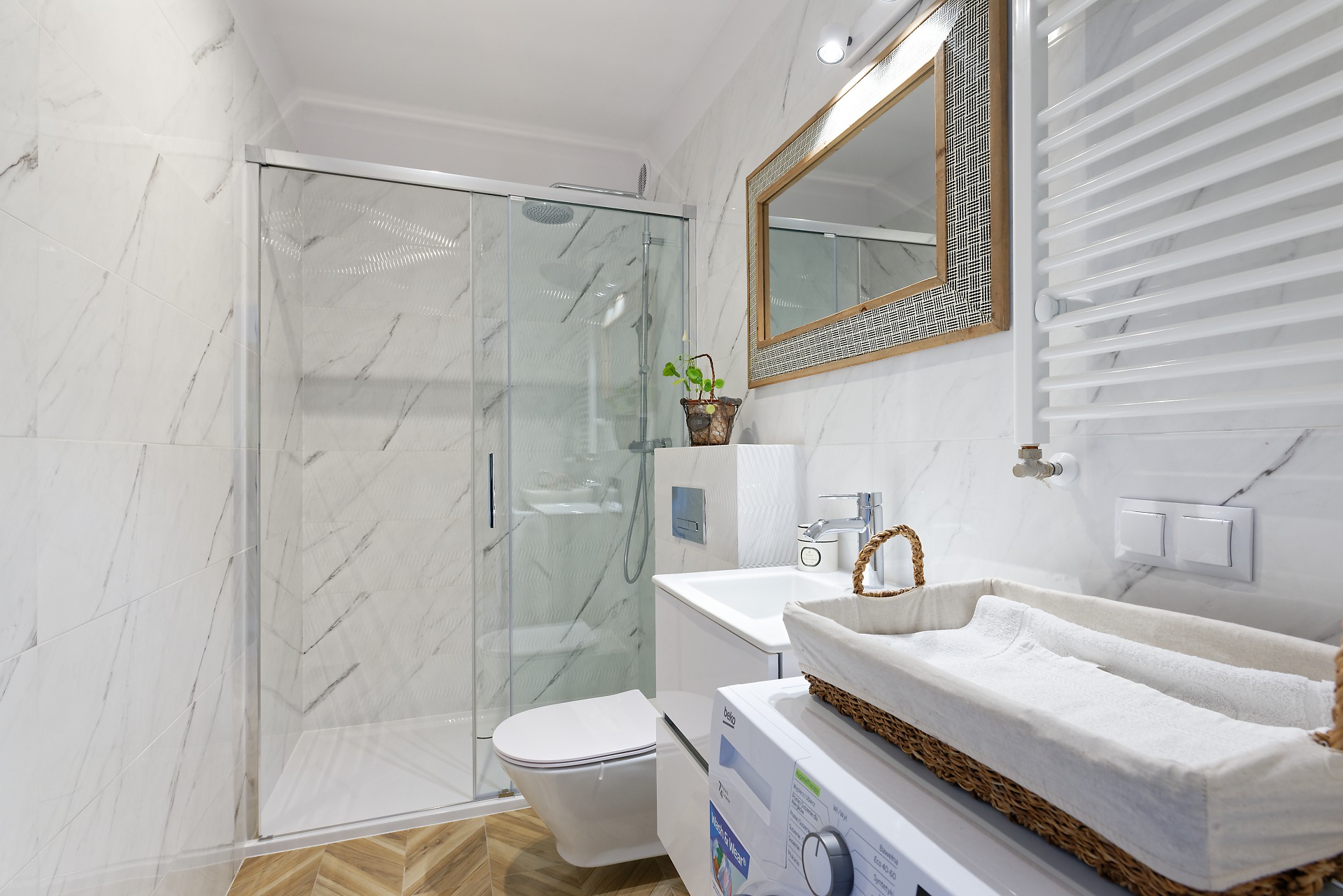 Nowoczesna łazienka w bieli z marmurowym wzorem na płytkach, kabina prysznicowa ze szklanymi drzwiami przesuwnymi, drewniane akcenty w ramie lustra i koszu na pranie.
