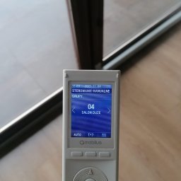 SERWIS OKIEN I DRZWI OKNO-SPEC RAFAŁ TURECKI - Biały pilot do sterowania roletami z wyświetlaczem wskazującym grupę 'Salon Duże' i datę 2021-11-04, na tle okna.