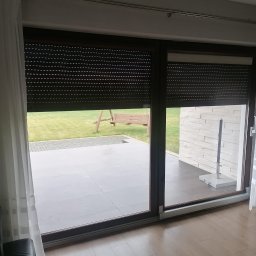 SERWIS OKIEN I DRZWI OKNO-SPEC RAFAŁ TURECKI - Zasłonięte brązowe rolety zewnętrzne na dużym oknie tarasowym, częściowo odsłonięte, z widokiem na ogród z huśtawką i zewnętrzny taras z jasną elewacją.