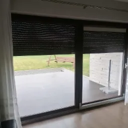 Zasłonięte brązowe rolety zewnętrzne na dużym oknie tarasowym, częściowo odsłonięte, z widokiem na ogród z huśtawką i zewnętrzny taras z jasną elewacją.