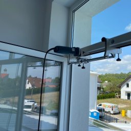 SERWIS OKIEN I DRZWI OKNO-SPEC RAFAŁ TURECKI - Narzędzie do naprawy okien zamocowane na ramie okiennej, widok z zewnątrz na domy i niebo.