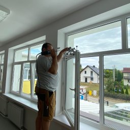 SERWIS OKIEN I DRZWI OKNO-SPEC RAFAŁ TURECKI - Mężczyzna w krótkich spodenkach montuje okno za pomocą specjalistycznego narzędzia; w tle widoczne domy jednorodzinne i drugi mężczyzna przy innym oknie.