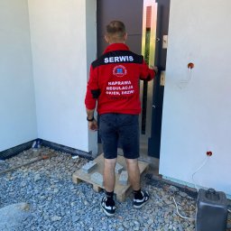 SERWIS OKIEN I DRZWI OKNO-SPEC RAFAŁ TURECKI - Mężczyzna w czerwonej kurtce serwisowej z napisem 'Naprawa Regulacja Okien, Drzwi' otwiera nowoczesne, grafitowe drzwi wejściowe do nowo wykończonego domu z elewacją w kolorze złamanej bieli...