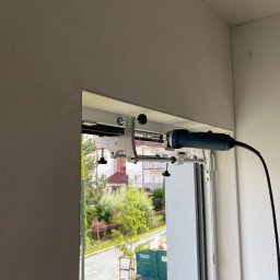 SERWIS OKIEN I DRZWI OKNO-SPEC RAFAŁ TURECKI - Montaż rolety dzień noc: frezarka zamocowana do ramy okiennej za pomocą specjalnego uchwytu, widok na zewnątrz przez okno, zieleń drzew i budynki w tle.