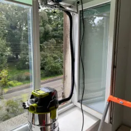 Frezarka z odkurzaczem przemysłowym zamontowana na ramie okiennej podczas prac renowacyjnych, widok z zewnątrz na zieleń.