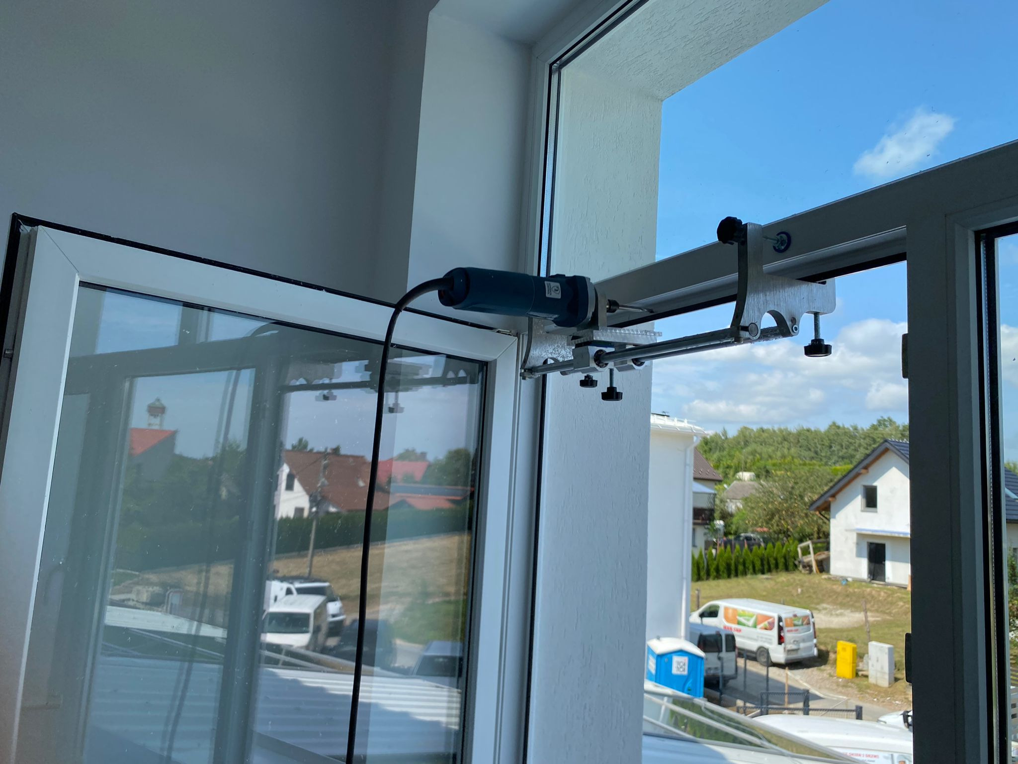 Narzędzie do naprawy okien zamocowane na ramie okiennej, widok z zewnątrz na domy i niebo.