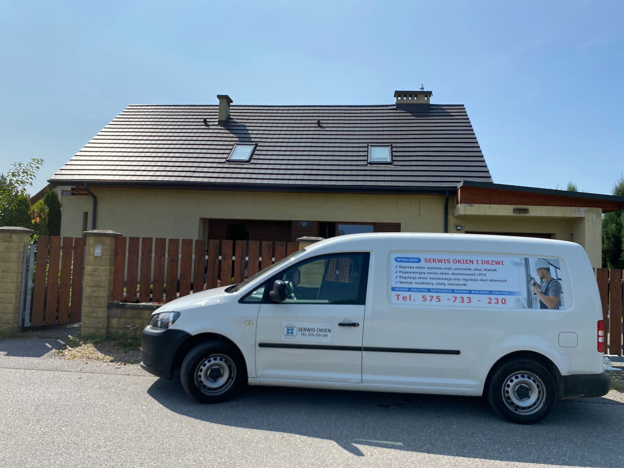 Biały samochód serwisowy z reklamą naprawy okien i drzwi zaparkowany przed domem jednorodzinnym z oknami dachowymi.