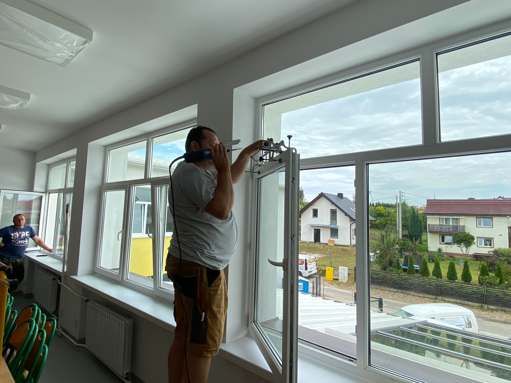 Mężczyzna w krótkich spodenkach montuje okno za pomocą specjalistycznego narzędzia; w tle widoczne domy jednorodzinne i drugi mężczyzna przy innym oknie.