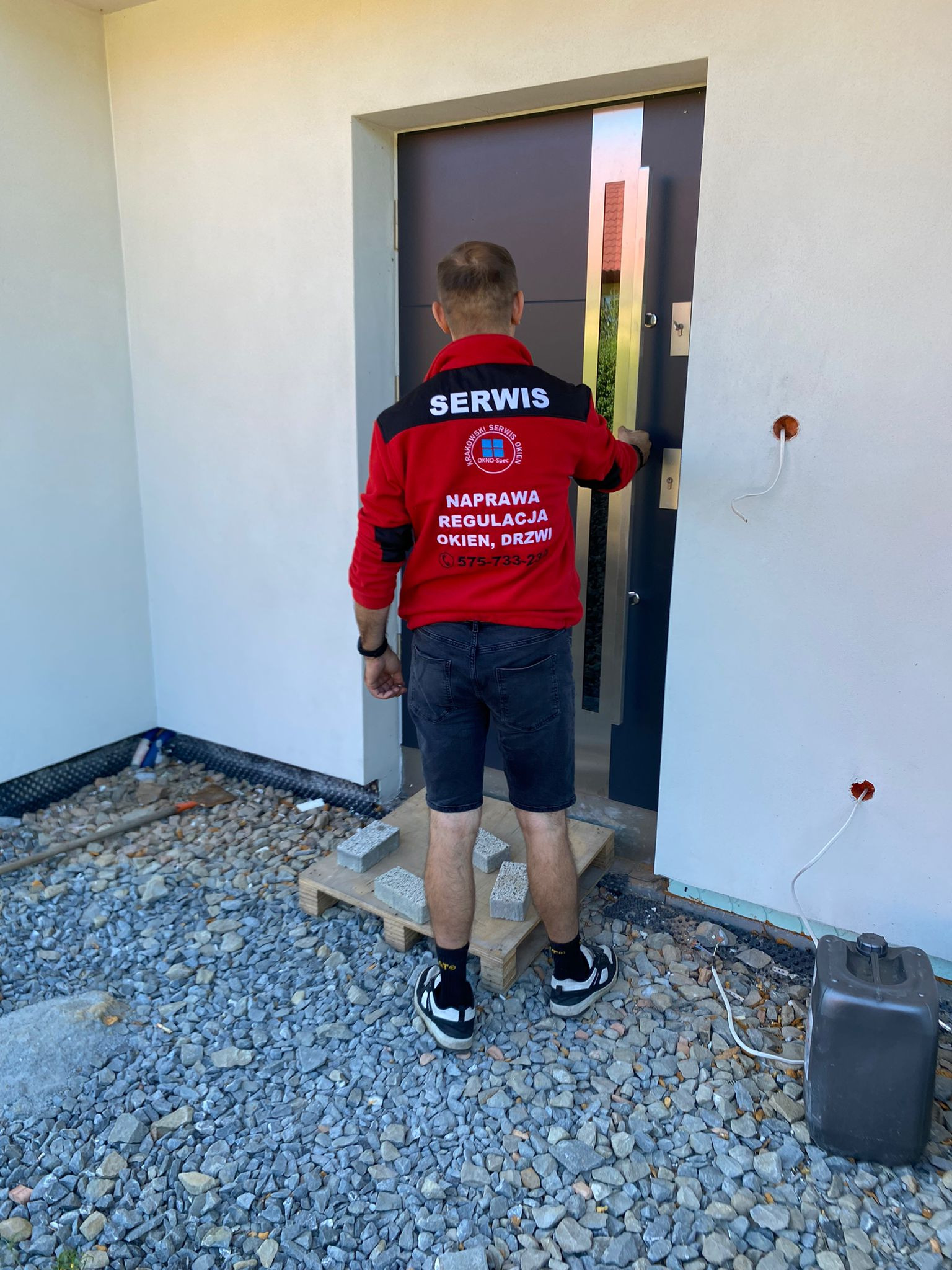 Mężczyzna w czerwonej kurtce serwisowej z napisem 'Naprawa Regulacja Okien, Drzwi' otwiera nowoczesne, grafitowe drzwi wejściowe do nowo wykończonego domu z elewacją w kolorze złamanej bieli...