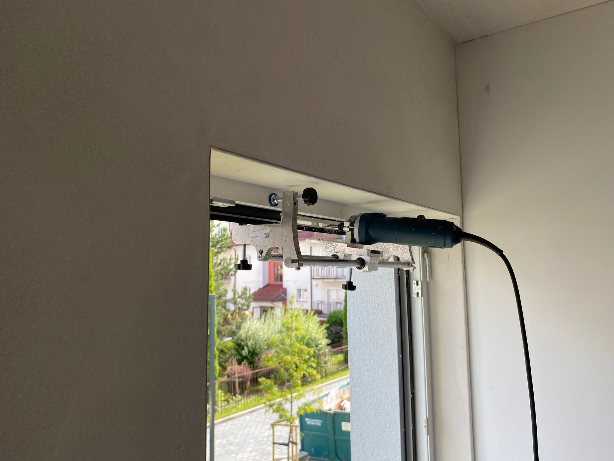 Montaż rolety dzień noc: frezarka zamocowana do ramy okiennej za pomocą specjalnego uchwytu, widok na zewnątrz przez okno, zieleń drzew i budynki w tle.