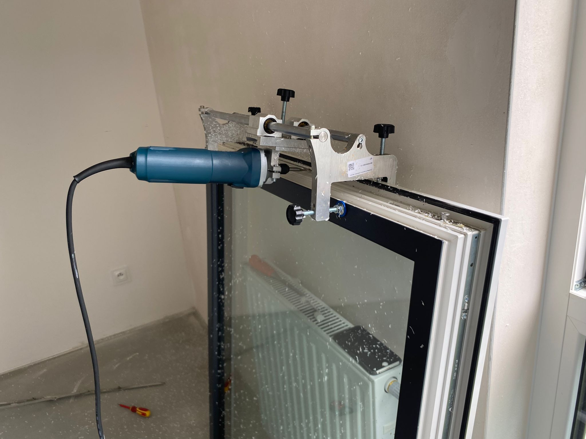 Frezarka przymocowana do ramy okiennej podczas obróbki, widoczne wióry i radiator za oknem.