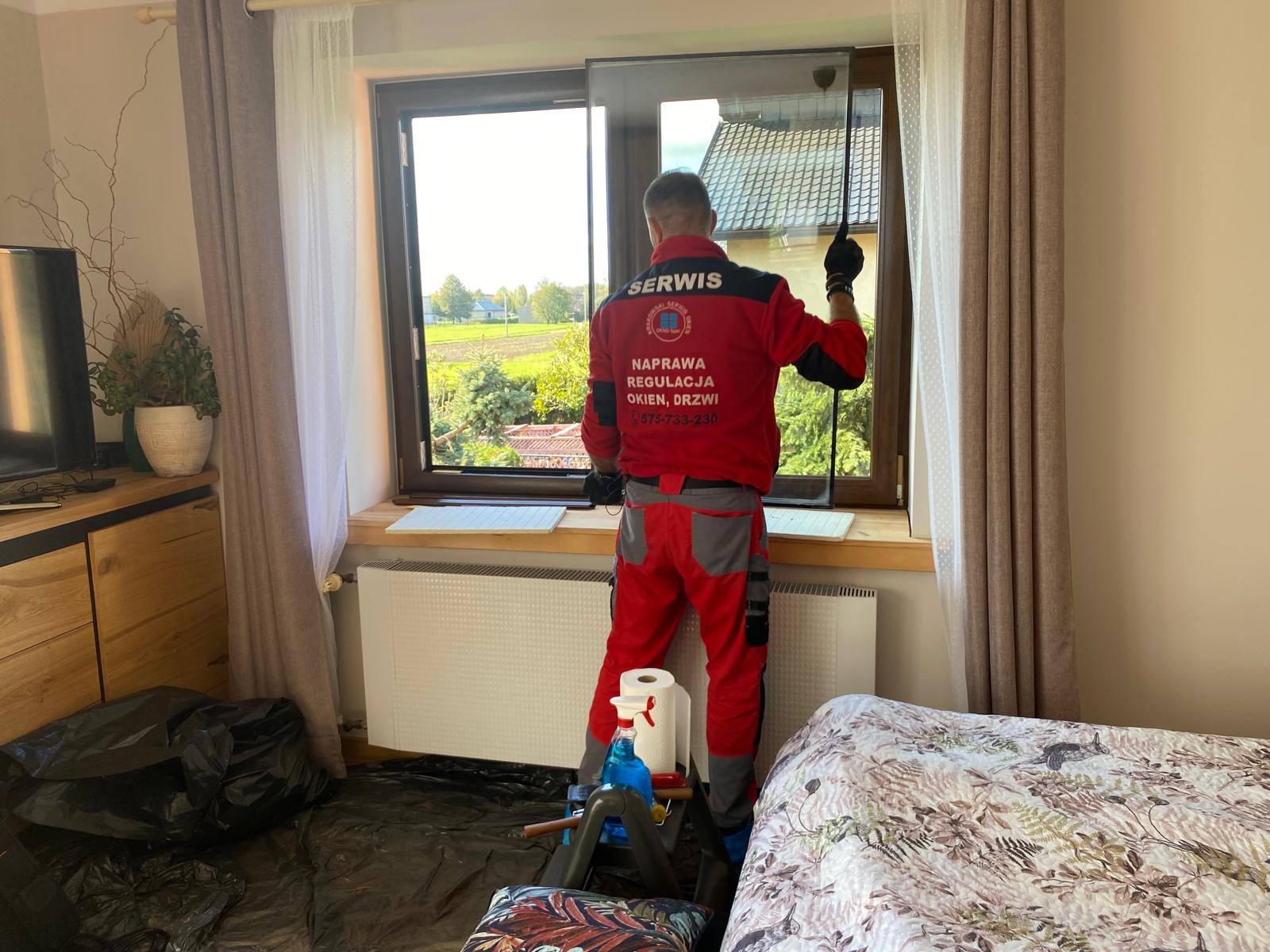 Mężczyzna w uniformie serwisowym montuje szybę w oknie z brązową ramą; widoczny krajobraz za oknem i zabezpieczone folią meble.