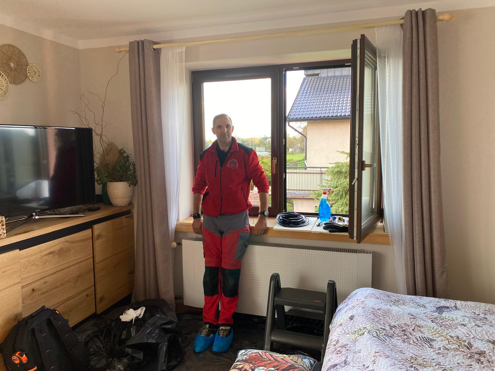 Mężczyzna w roboczym stroju i ochraniaczach na butach stoi przy otwartym oknie, obok leżą narzędzia i akcesoria potrzebne do naprawy, na podłodze folia ochronna.