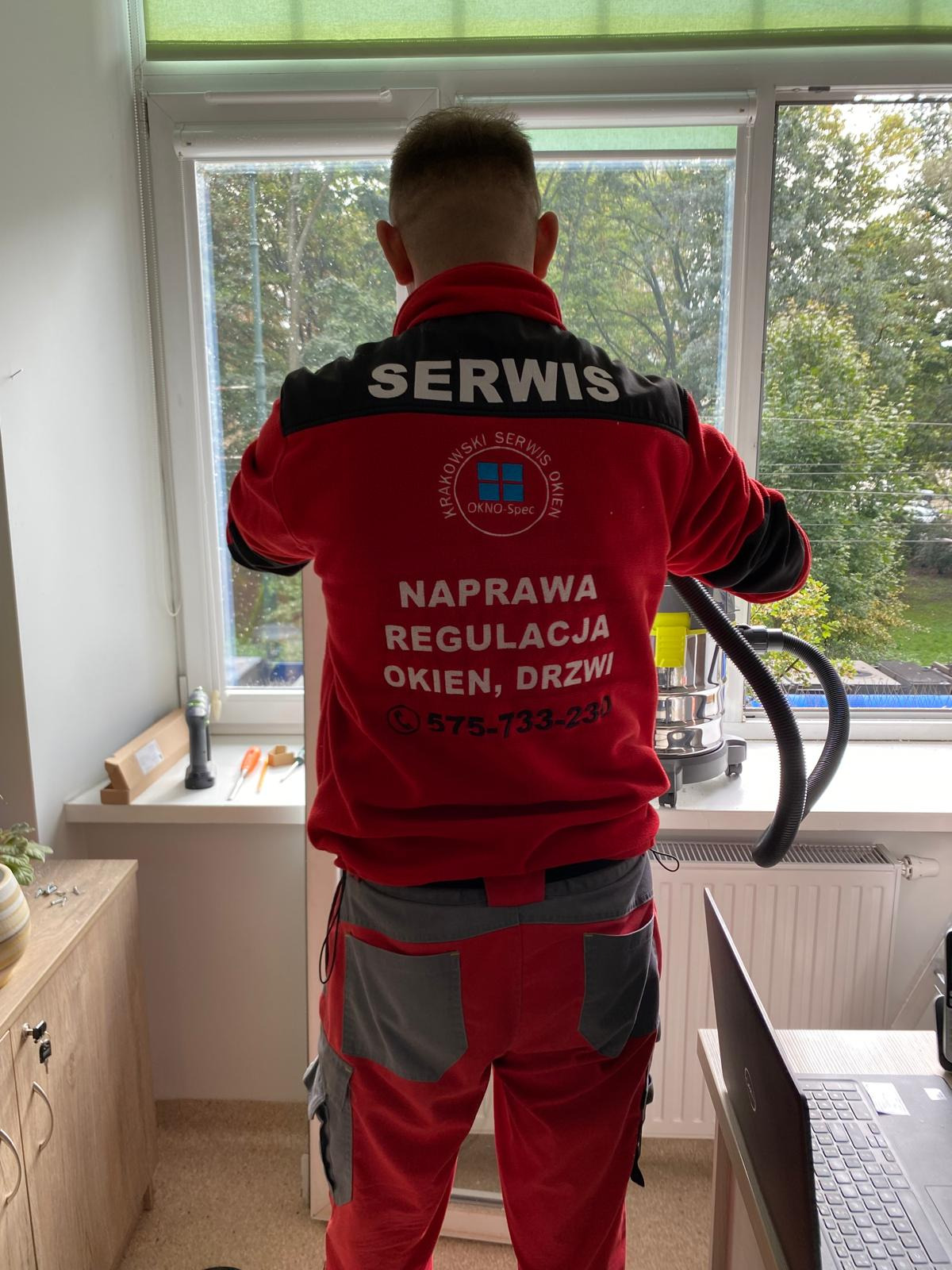 Pracownik serwisu okien w czerwonej bluzie z napisem firmy, używa odkurzacza do czyszczenia ramy okiennej po naprawie, widok z tyłu, na parapecie leżą narzędzia.