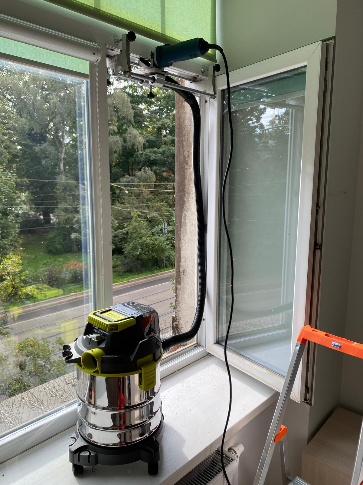 Frezarka z odkurzaczem przemysłowym zamontowana na ramie okiennej podczas prac renowacyjnych, widok z zewnątrz na zieleń.