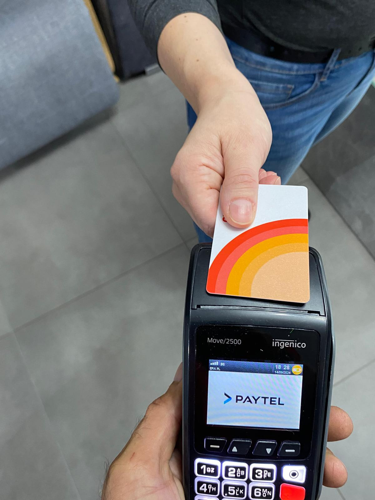Kobieta wkłada kartę płatniczą z tęczowym motywem do terminala płatniczego Ingenico Move/2500 z widocznym logo Paytel na ekranie.