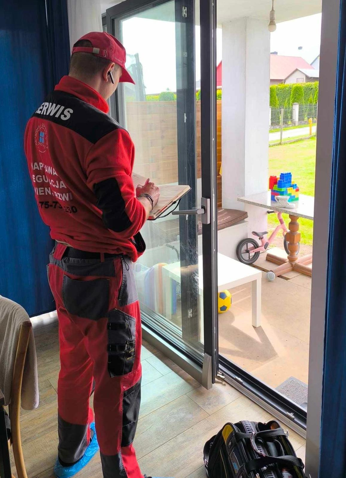 Mężczyzna w czerwonym kombinezonie serwisowym z logo firmy, stojący tyłem do kamery, trzymający clipboard i oglądający przesuwne okno tarasowe z widokiem na podwórko z zabawkami.