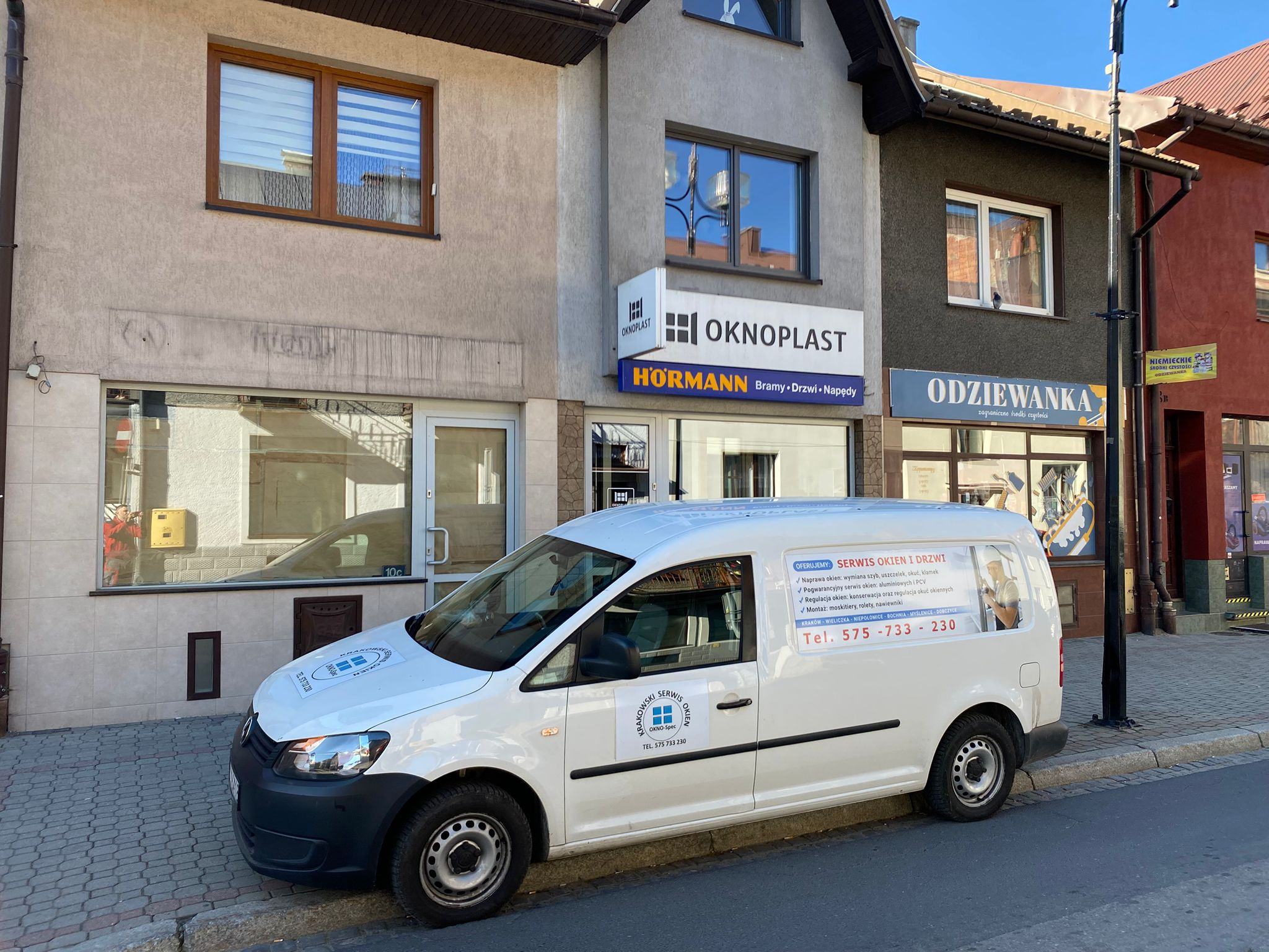 Biały samochód dostawczy z reklamą serwisu okien i drzwi parkuje na tle budynków z oknami różnych typów. Na samochodzie widoczne logo firmy i numer telefonu.