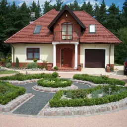 Ogród z krzewami uformowanymi w geometryczne wzory, obrzeża z kostki, podjazd z jasnej kostki brukowej prowadzący do domu z czerwonym dachem i garażem.