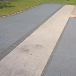 Ułożona nawierzchnia z kostki betonowej w dwóch odcieniach szarości i beżu, oddzielona pasem drobnego kruszywa, prowadząca do schodów wejściowych budynku.