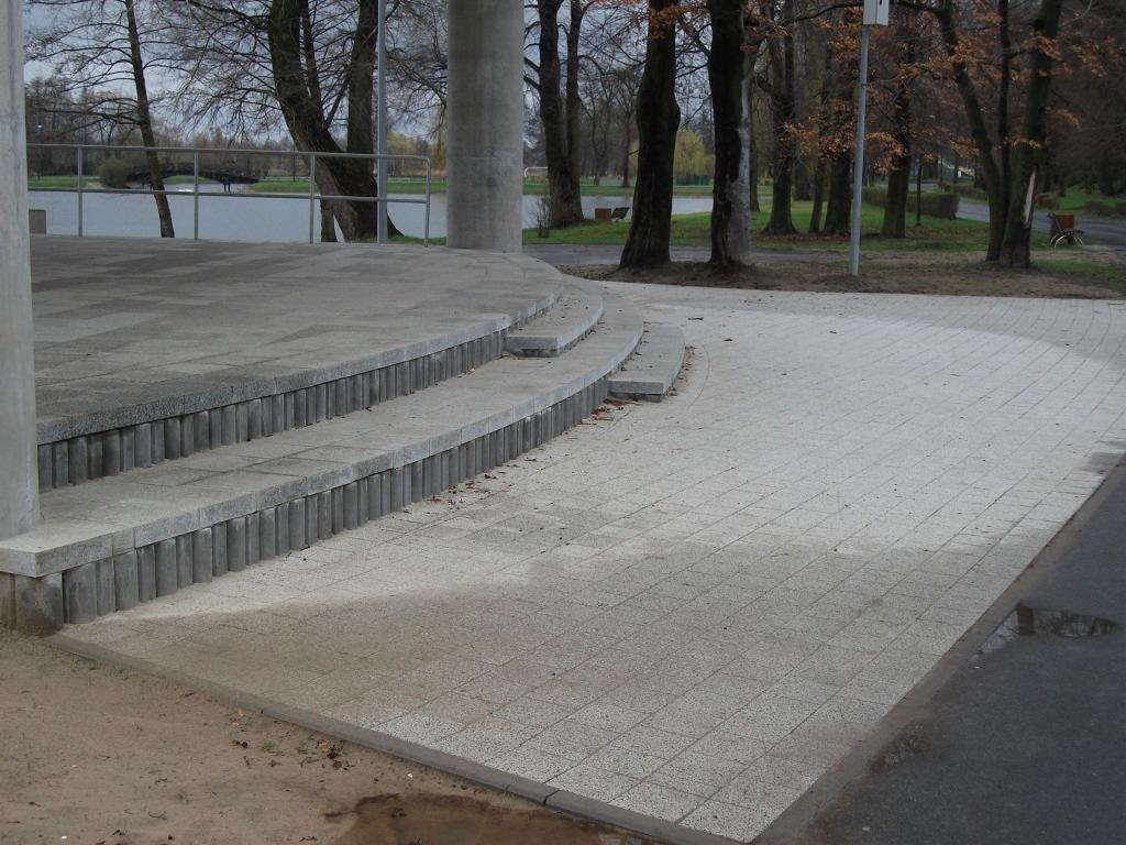 Szara kostka brukowa tworzy ścieżkę w parku, prowadzącą do podwyższonej platformy z betonowymi schodami. Widok na drzewa i jezioro w tle, jesienna sceneria.