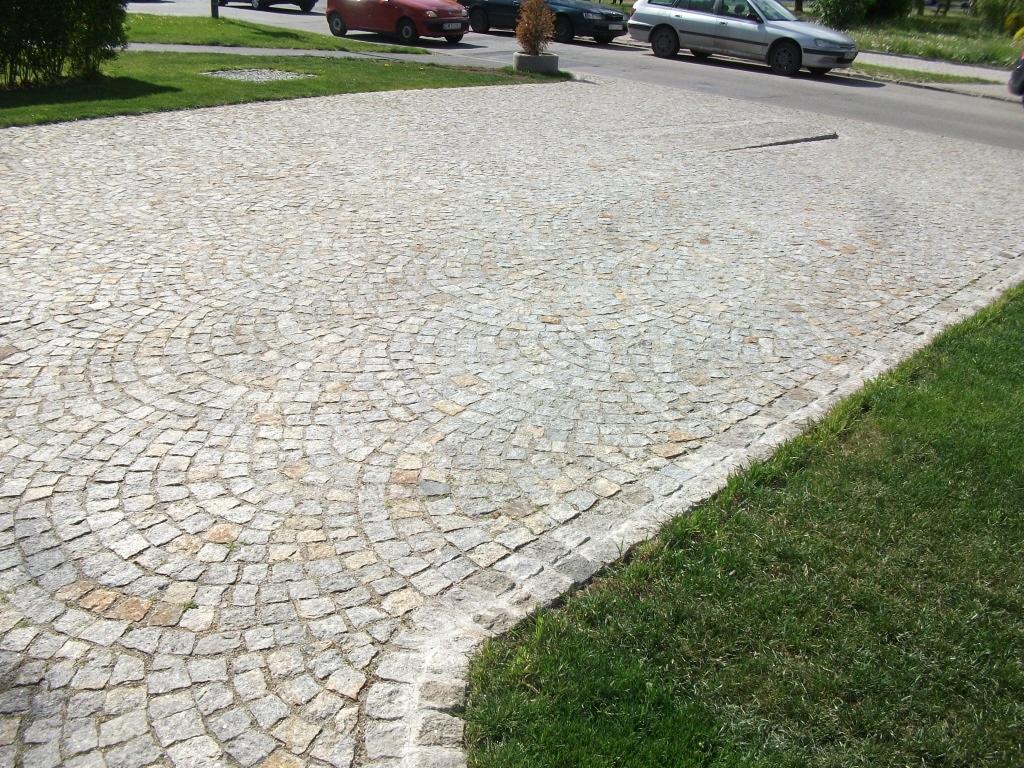 Ułożona, jasnoszara kostka granitowa tworzy okrągły wzór na parkingu, z zielonym trawnikiem po prawej stronie i zaparkowanymi autami w tle.
