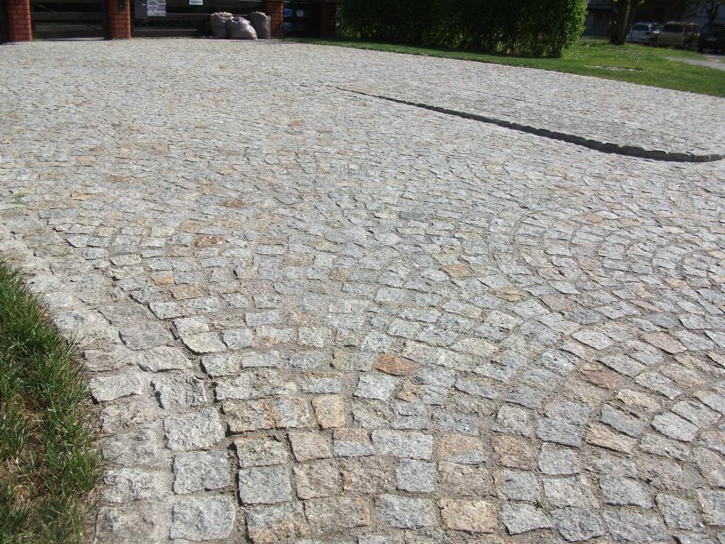 Granitowa nawierzchnia z elementami ułożonymi w łukowaty wzór, obrzeżem i fragmentem trawnika. Widok na podjazd z workami w tle.